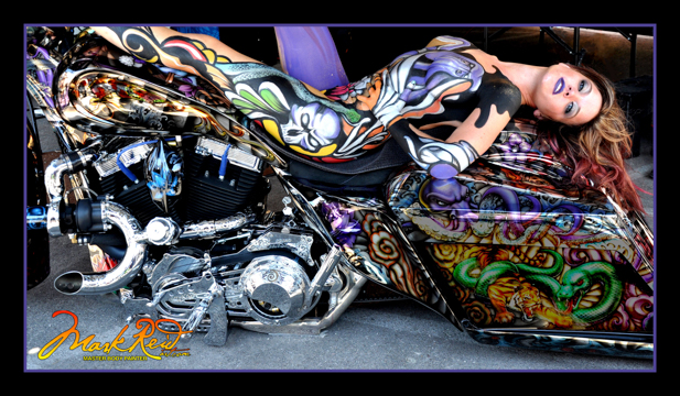 brunette woman with her entire body painted in a very elaborate colorful design that matches the motorcycle she is laying on her back on with her feet on on the handle bars